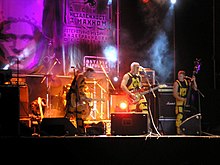 Photograph of a rock band playing on a stage, with a large image of Nestor Makhno behind them