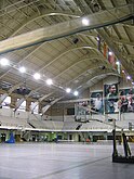 A broad view of the gym's interior showing the lofty rafters. The gym's walls are lined with about ten rows of elevated bleachers, and the back wall features photos of Michigan State athletes. Several basketball hoops line the grey playing surface.