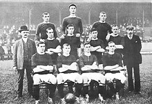 A black-and-white photo of a football team. The players are on three levels, with four on the bottom row, four on the middle row and three on the top row. The player sitting second from the left on the bottom row has a ball at his feet, and the player in the middle of the top row is wearing a flat cap.