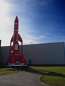 A model of a rocket-shaped spacecraft with three engine nacelles around its cylindrical main body. Photographed standing upright in front of a building.