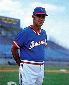 A man in a blue cap with a white "N" on the front, a blue baseball jersey with "Sounds" across the chest in white and red, and white pants stands on a baseball field with his hands behind his back.