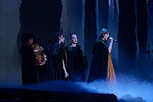 Taylor Swift, dressed in a burnt orange dress and black cloak, singing on a microphone with backup dancers also dressed in black cloaks