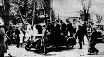 A black-and-white photograph showing several hooded Ku Klux Klan members, one of whom is carrying an American flag. A few policemen are near the Klansmen, and more officers stand in a nearby car. To the right, a sashed Klansman is elevated. Civilians are intermixed and looking upon the scene. The letters "PEA" appear at bottom right.