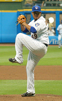 A man in a white baseball uniform with blue stripes and a blue cap standing on the mound in the midst of pitching the ball
