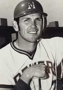 A man in a light baseball uniform and a dark batting helmet with an "M" on the center resting his hand on the knob of a baseball bat