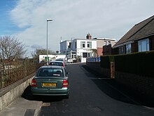 Under a partly cloudy sky, a concrete roadway recedes into the distance where a two-storey, flat-roofed, white stone building can be seen. A green car is parked behind a row of three cars. On the left is a low concrete wall with iron railings and a lamp post. On the right are a hedge and wall surrounding a bungalow.
