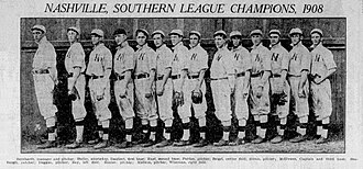 A black and white photograph of fourteen men standing in front of a fence wearing white baseball uniforms with a dark "N" on the chests and light caps with dark brims; some are wearing fielding gloves.