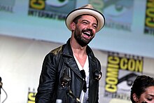 A brunette man wearing a fedora, white t-shirt, and black jacket laughs as he looks off to the crowd
