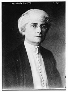 A white woman with white hair and dark eyebrows, wearing eyeglasses, a high-collared white blouse with many fabric-covered buttons down the front, and a dark jacket