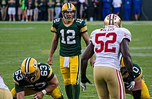 A photo of Aaron Rodgers lining up to snap the ball against the 49ers. Rodgers is viewed straight on, while Patrick Willis is viewed from behind. Other Packers lineman are in the photo.
