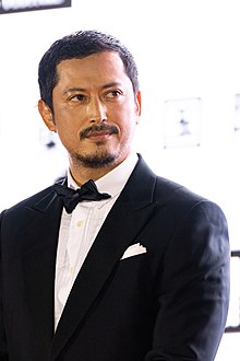 Ikeuchi at press event wearing tuxedo and looking to right of camera