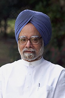 A portrait photograph of a bespectacled Indian man with a dark grey beard, a blue turban, and a white button-down shirt standing before a natural backdrop of trees. A pen is in his pocket.