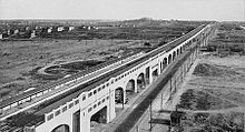 Overhead shot of a long, concrete train causeway. On either side are dirt roads and empty lots stretching to the horizon.