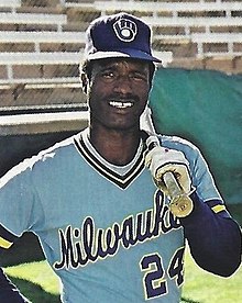A man in a powder blue baseball jersey with "Milwaukee" written across the chest and a blue cap resting a baseball bat on his shoulder