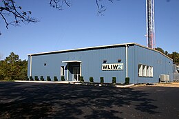 A one-story blue metal building with a W L I W logo next to the base of a tall broadcasting tower. In comparison to the prior photo, the building has been reshaped in the front and is larger, with new landscaping.