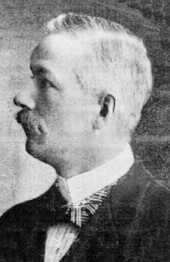 Black and white photo of a middle-aged man facing toward the photo's left, with a large moustache, wearing a dark suitcoat and a white dress shirt