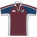 A maroon coloured rugby jersey; maroon on the shoulders and top of the sleeves with the mid three-quarters of the sleeves in blue and the underside being grey. A large white collar and thin white lines on the sleeves.
