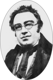 Round-faced white man, clean shaven with unruly dark hair and round spectacles
