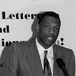 A black-and-white photo of a black man wearing a tie and a suit standing behind a microphone