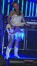 Ginger Reyes—a Caucasian woman in her late 20s with long brunette hair—plays bass guitar on stage wearing a white dress and white cowboy boots.