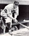 A man in an off-white jersey and pants, a baseball cap with the letter "B" in the center, and dark baseball socks leans forward holding a baseball bat with both hands.
