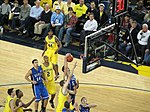 Two opposing basketball teams on a basketball court fight for a rebound. One team is in maize uniforms with the word Michigan on the front and names on the back and the other is light blue with the word Duke on the front and names on the back.