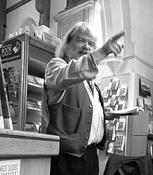 photo of a rotund bearded jolly man pointing to the audience with one hand while reading from the book of his poetry with his other.