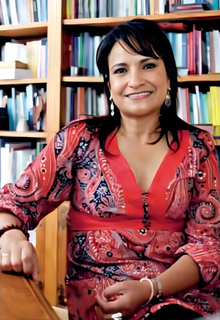 Official portrait of Marcela Revollo; sat at a table, bookshelf in the background.