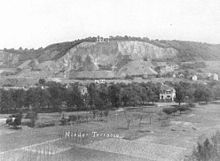 A black and white photo of a hillside quarry amidst countryside, showing a site in the middle of the quarry marked by a small white arrow.