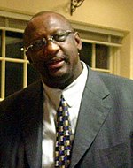 A black person, wearing a gray suit, a tie and glasses, is looking to the front.