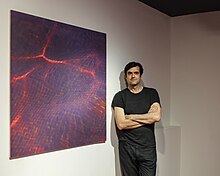 A man with a serious expression and dressed in all black leaning against a wall arms crossed over his chest. Next to him is his photo, Red Interlude, an abstract image of a red pool.