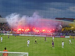 A football stadium full of supporters, which are using flares and smoke bombs.
