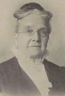 An older white woman, grey hair arranged in a bun on top of her head; she is wearing eyeglasses and a blouse with high frilled collar with a dark jacket