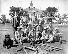 Group photograph of baseball players wearing 1910s-era uniforms, standing, kneeling, or laying on the ground behind a collection of baseball bats