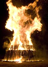 Large flames engulf the top layer of a circular three-tier structure of logs; the logs are vertical to the ground.