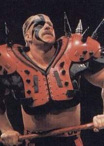 A man in face-paint wearing spiked shoulder pads looks off into the distance
