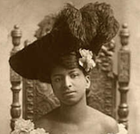 sepiatone headshot of a young woman sitting in a wooden chair, she is wearing a large black hat with elaborate feathers in it