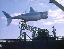 A large model shark is hoisted by a crane as two men watch it.