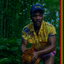 Glover, wearing a bright yellow shirt, short purple shorts and blue SPAM cap, crouches in the jungle holding a chicken and looking concerned.