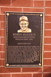 A dark bronze plaque with a man's face at the top and gold writing beneath, mounted on a brick wall