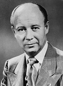 A black-and-white portrait photograph of a mostly bald middle-aged man in a dapper suit.