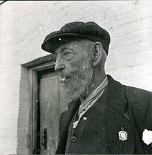 Bearded old man outside a rustic door wearing a flat cap and smoking a roll-up.