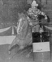 Black and white photograph of a woman in a cowboy hat and large print shirt riding a horse in a barrel racing competition.