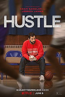 A downcast bearded man in a red sweater sitting at the edge of a basketball court. Players are a blur in the foreground.