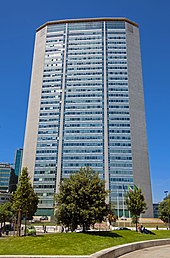 A narrow, unornamented skyscraper with blue-green glass windows in the middle and a tapered metallic skin on the sides rising above some trees at ground level against a blue sky