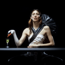 Polachek seated at a table with makeup and a costume making her arm resemble a swan