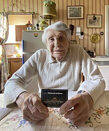 Image of an elderly woman, holding a small card, in a woollen jumper