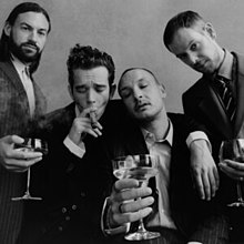 In a black-and-white image, three of the four band members of the 1975 are seen holding wine glasses while the lead singer, Matty Healy (centre left) smokes a cigar.