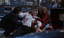 A man in a white shirts collapses into the arms of a young man in a suit and a woman dressed in red.