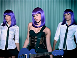 Shakira appears in the middle wearing a purple wig and a black corset while holding a guitar on her shoulders. Behind her, two dancers appear wearing white shirts, black ties, miniskirts and purple wigs as well.